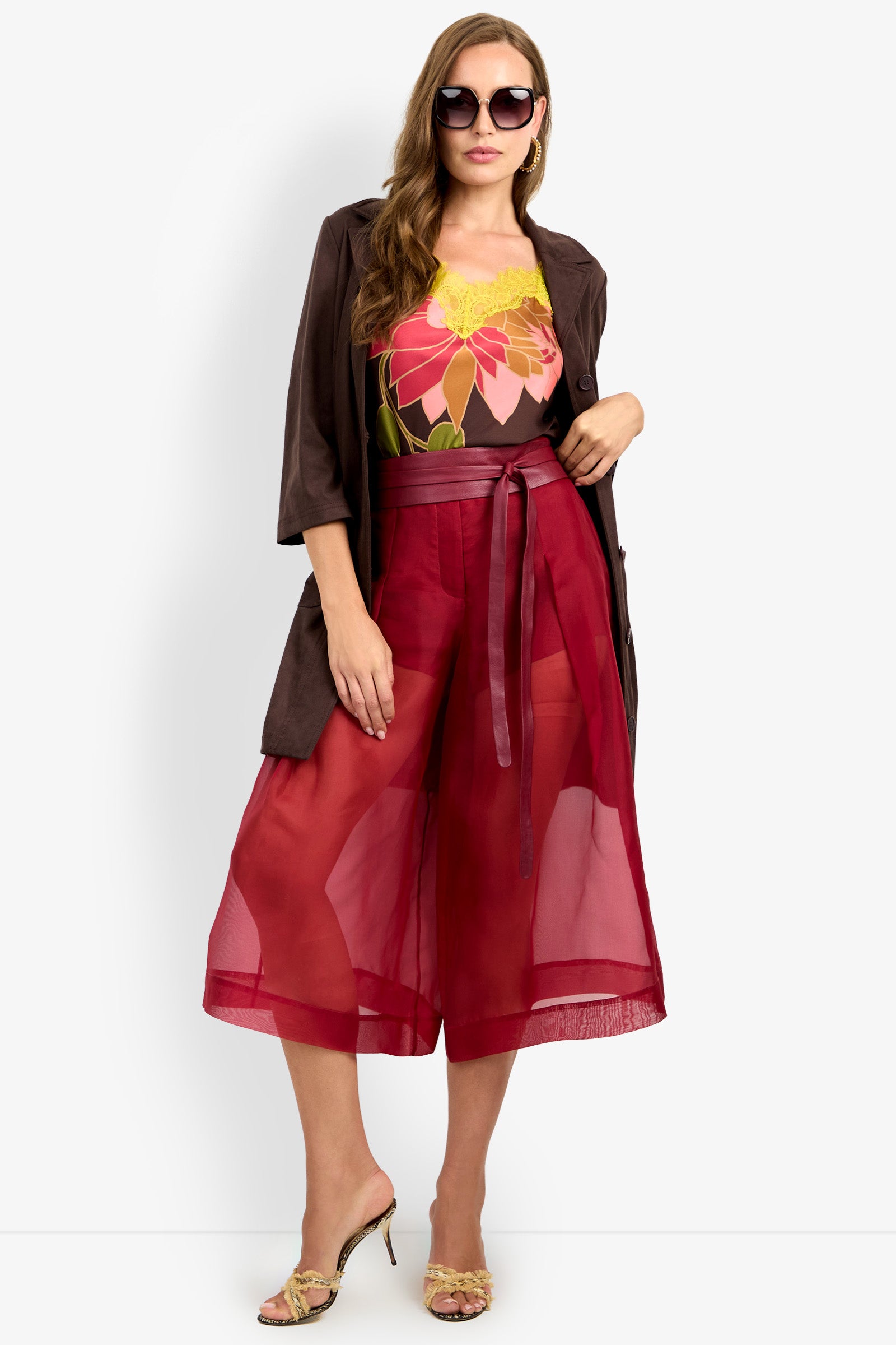 A woman stands against a plain white background wearing sunglasses, a brown jacket, a floral top, and the SIENA PANT—sheer red culottes with a sash belt—paired with open-toe heels.