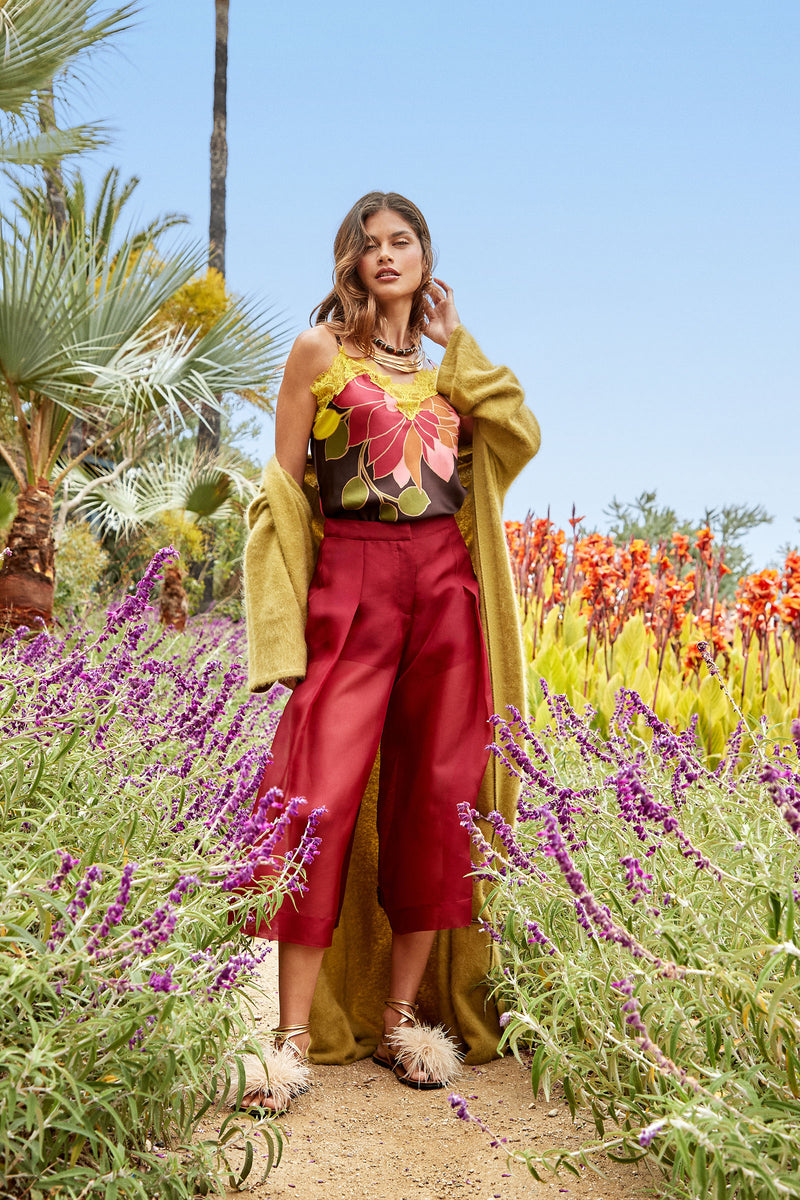A woman stands on a garden path surrounded by purple and orange flowers, wearing the Siena Pant, a floral top, fuzzy sandals, and a long yellow-green cardigan under a clear blue sky.