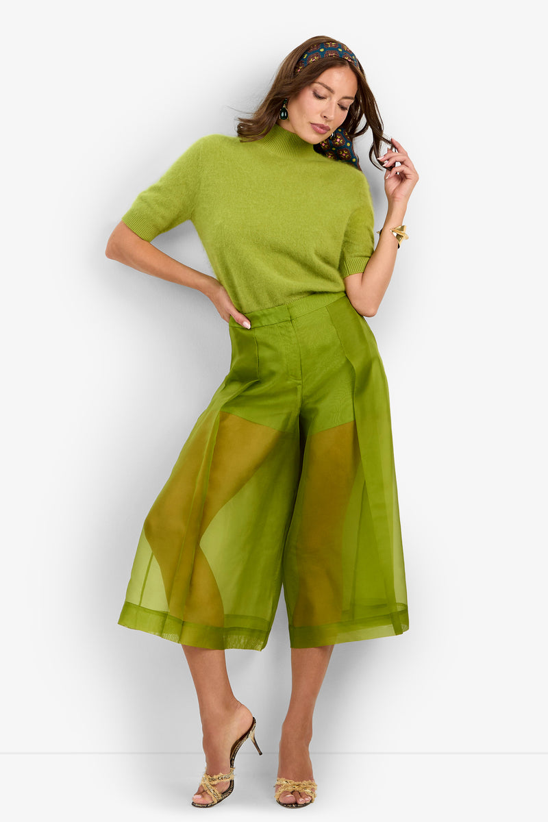 A woman stands against a white background in heels, wearing a light green short-sleeve sweater and the Siena Pant—sheer, wide-leg green trousers—with one hand on her head and the other at her waist.