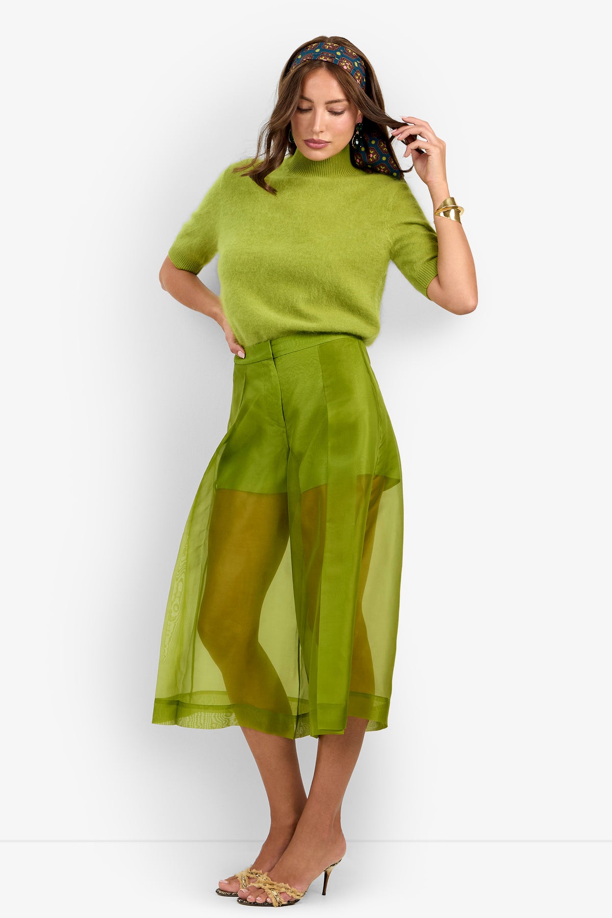 Woman wearing a fuzzy green short-sleeve top, sheer green midi skirt, floral headband, gold bracelet, open-toe heels, and the Siena Pant, posing with one hand on her head and the other at her side.