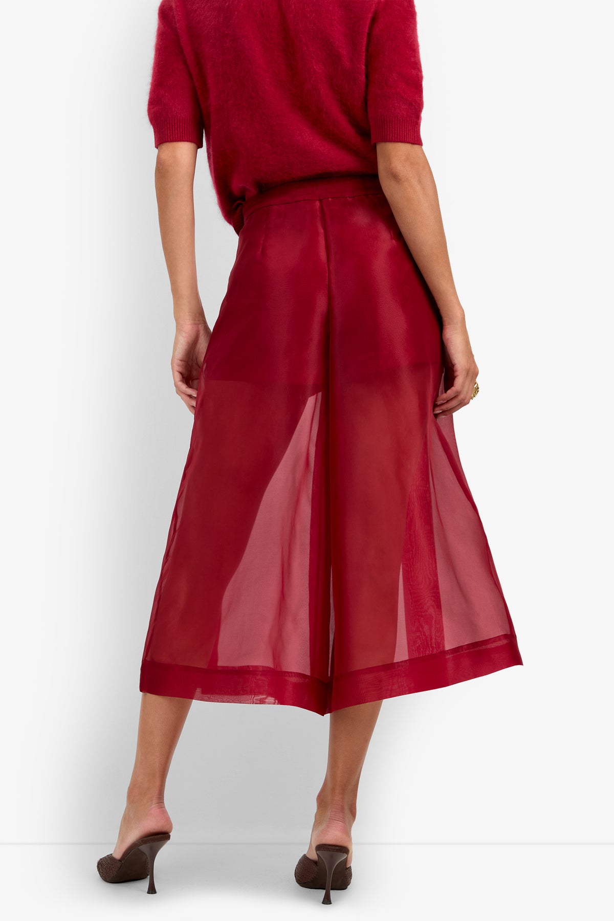 A woman stands with her back to the camera, wearing a dark red sweater, the Siena Pant in a sheer wide-leg red style, and brown heeled mules against a plain white background.