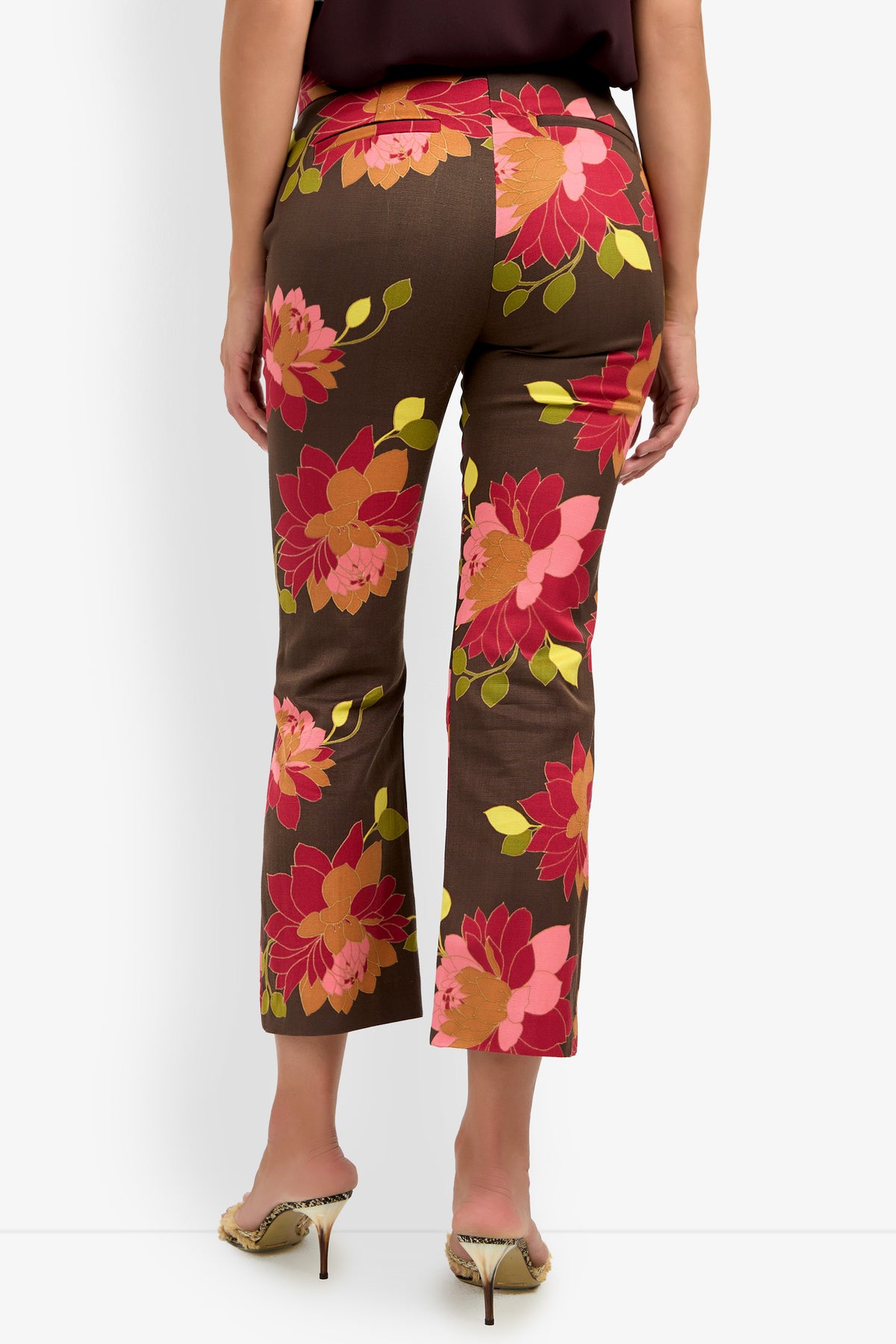 A woman is seen from the back wearing the LULU PANT, featuring a large pink, red, and yellow floral pattern on a brown background. She stands in open-heeled shoes with a small heel, with only her upper body partially visible.