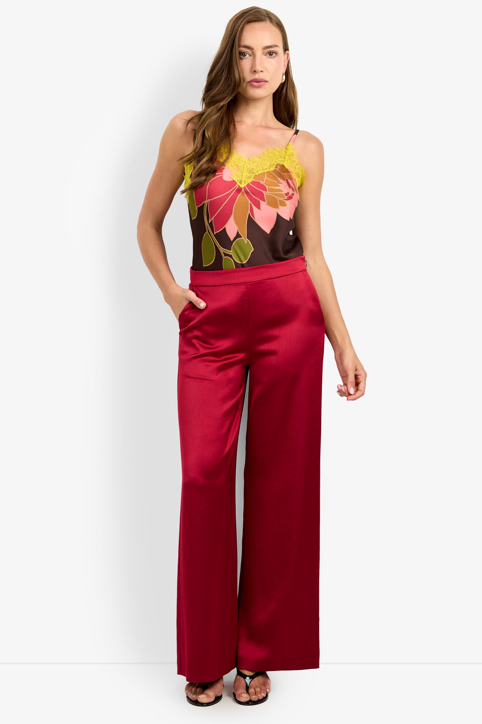 A woman stands against a plain background with one hand in the pocket of her INES PANT, wide-leg and shiny red, paired with a sleeveless floral top featuring yellow lace accents.