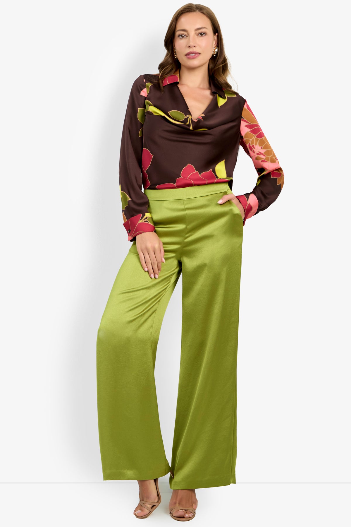 A woman wears the INES PANT—wide-leg, light green satin pants—paired with a brown floral blouse featuring a cowl neckline, standing against a white background with one hand in her pocket and one leg crossed.