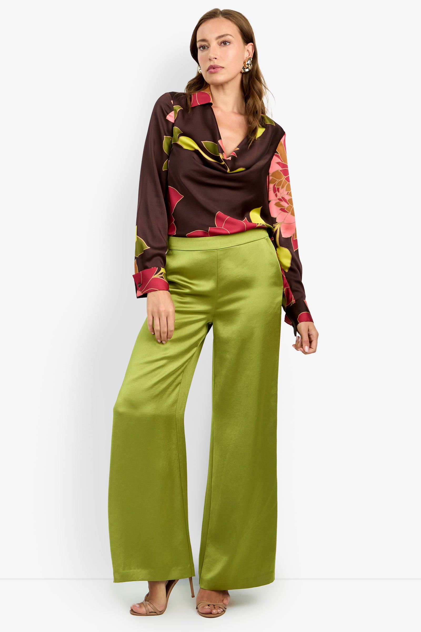 A woman stands in heels against a plain white background, wearing wide-leg, shiny green INES PANTS paired with a long-sleeve, floral-patterned brown blouse.