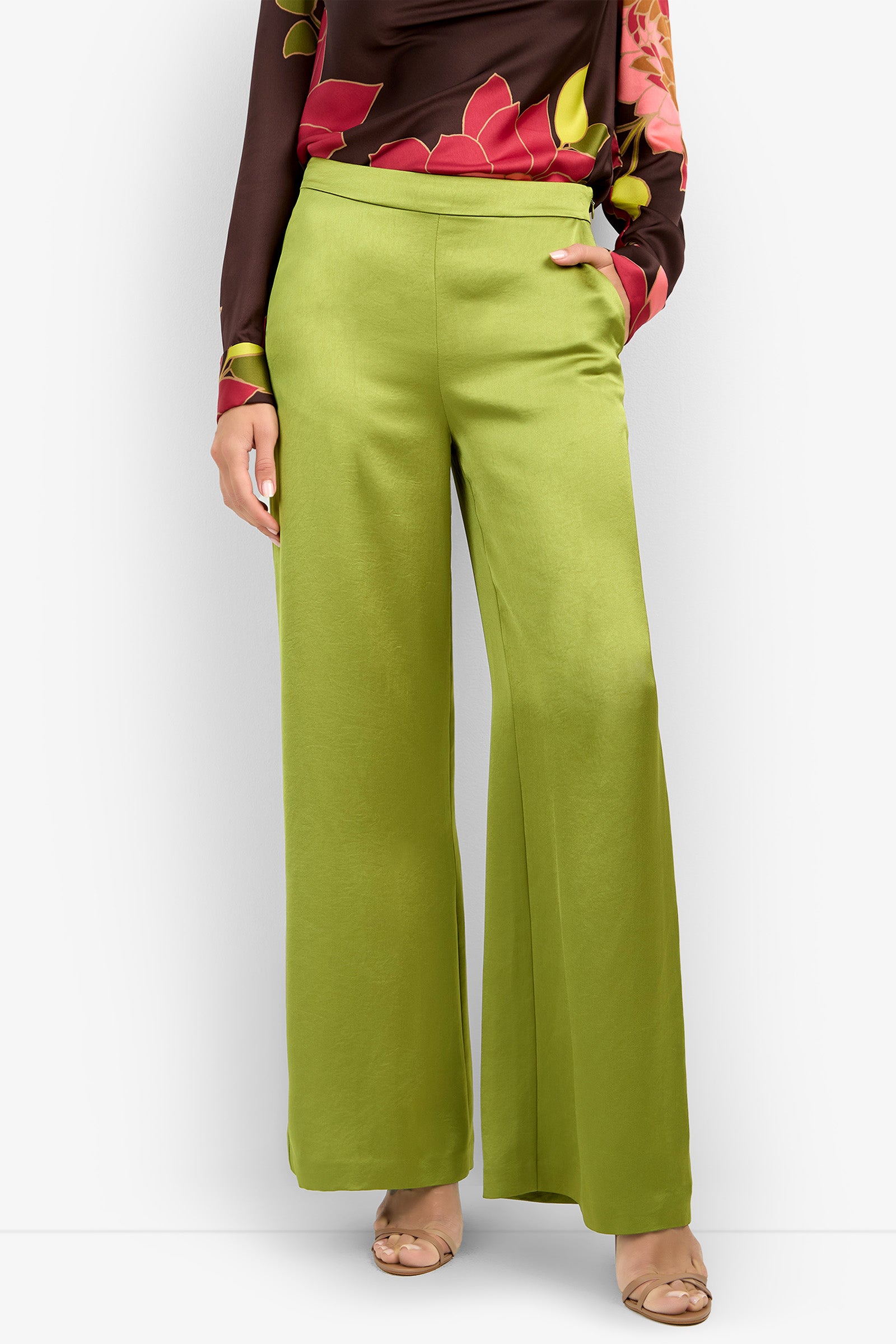 A person wearing the INES PANT—wide-leg, high-waisted green pants—paired with a long-sleeve brown top featuring a red and pink floral pattern and beige open-toe heels, standing against a white background.