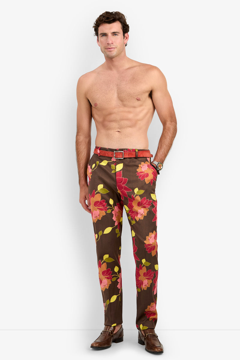 A shirtless man wearing the Clyde Slim Trouser—brown pants featuring large red and pink floral prints—with a matching belt, brown dress shoes, and a watch, stands against a plain white background.