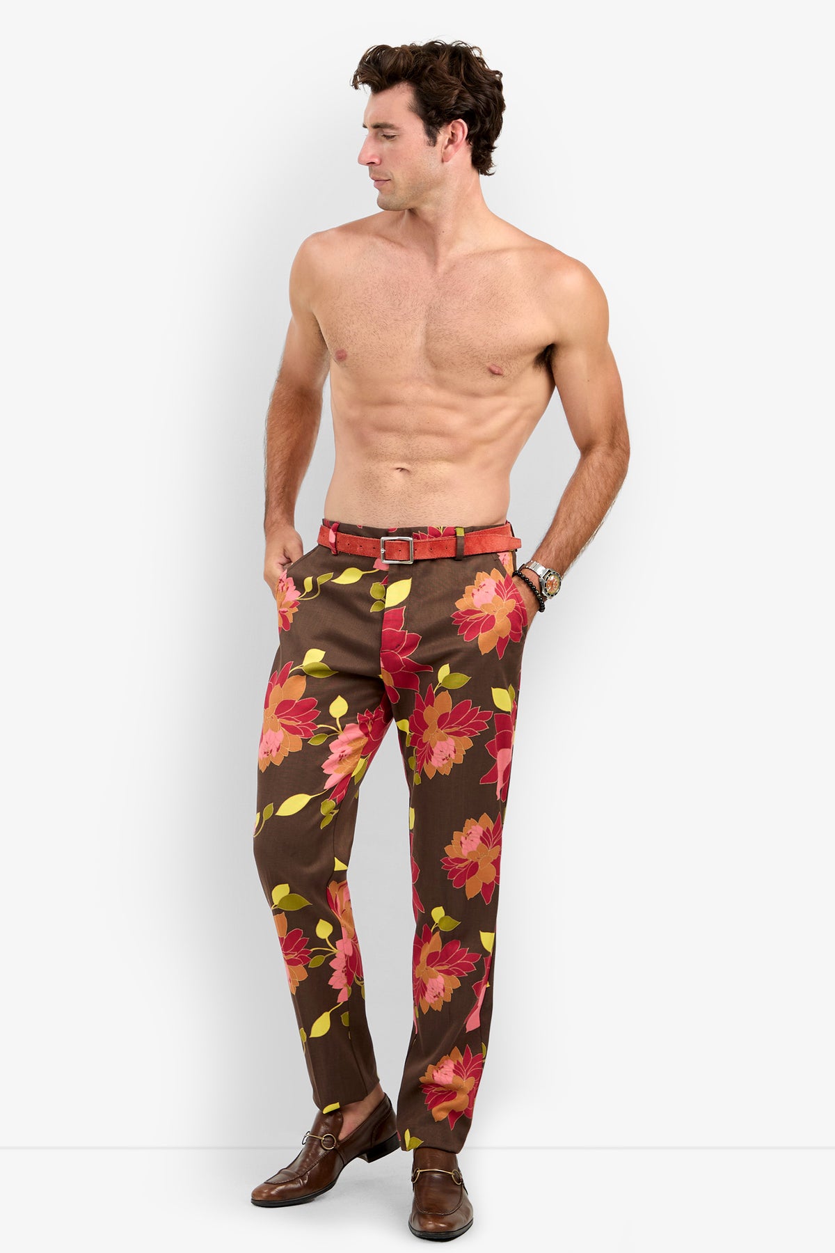 A shirtless man stands with his hands in his pockets, wearing the Clyde Slim Trouser—brown pants featuring a bold red and pink floral pattern—paired with brown shoes, a red belt, and a wristwatch. He looks to his left against a plain background.