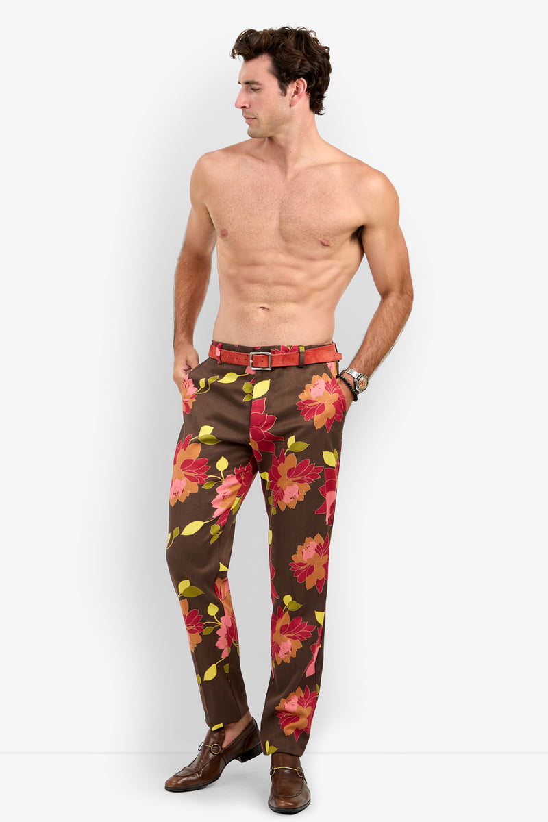 A shirtless man stands with his hands in his pockets, wearing the Clyde Slim Trouser—brown pants featuring a bold red and pink floral pattern—paired with brown shoes, a red belt, and a wristwatch. He looks to his left against a plain background.