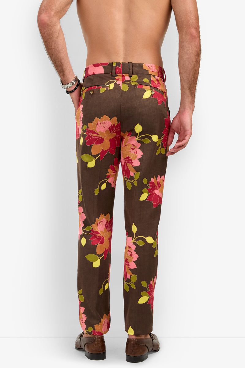 A shirtless man, seen from the back, models the Clyde Slim Trouser featuring a bold pink and yellow floral print over brown fabric. He accessorizes with a brown belt, bracelet, and matching brown dress shoes, standing against a plain white background.
