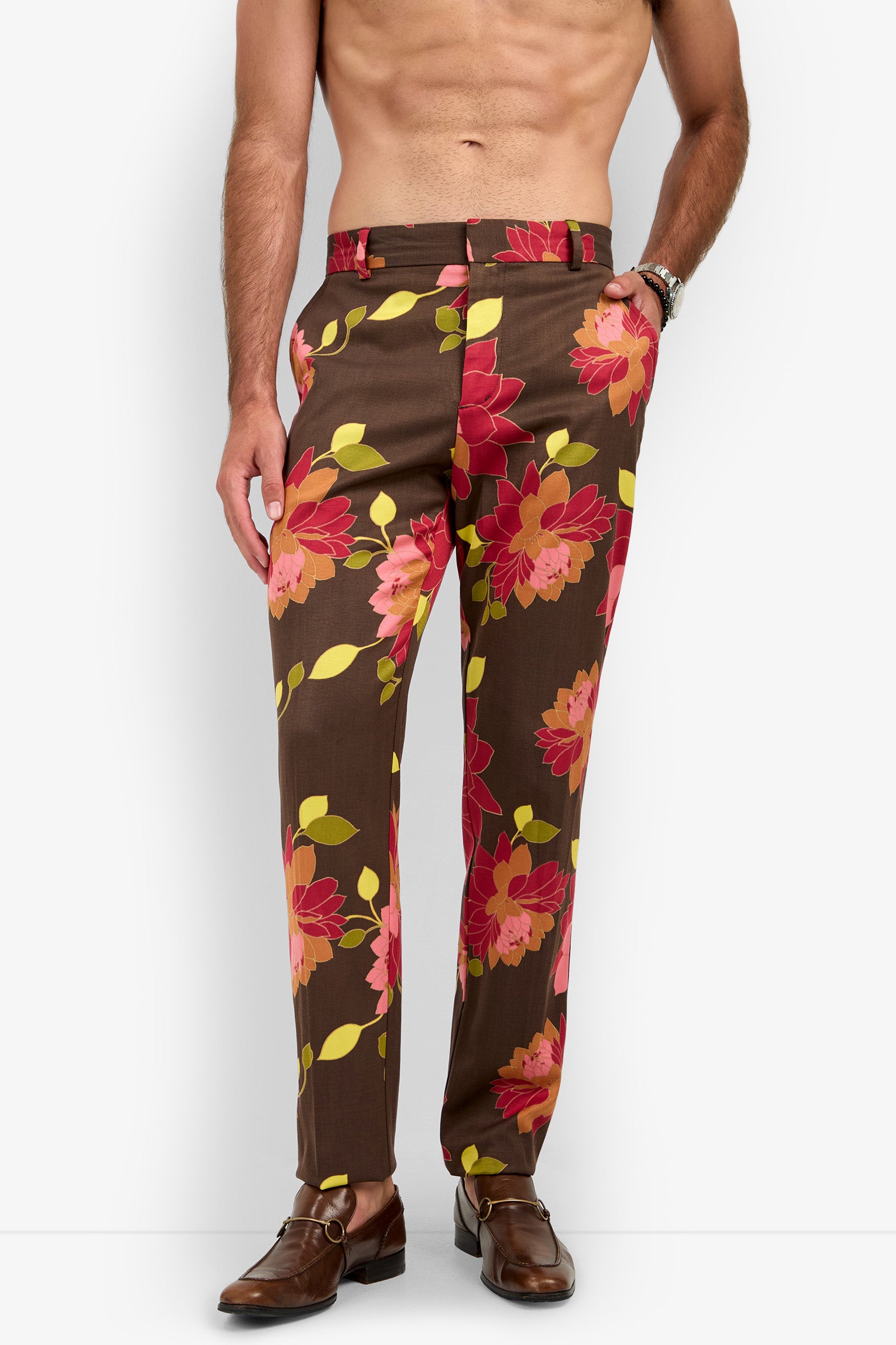 A shirtless man wears the “Clyde Slim Trouser,” featuring bold red, pink, and yellow floral prints on brown fabric, paired with brown loafers and a watch. His head is not visible in the image.