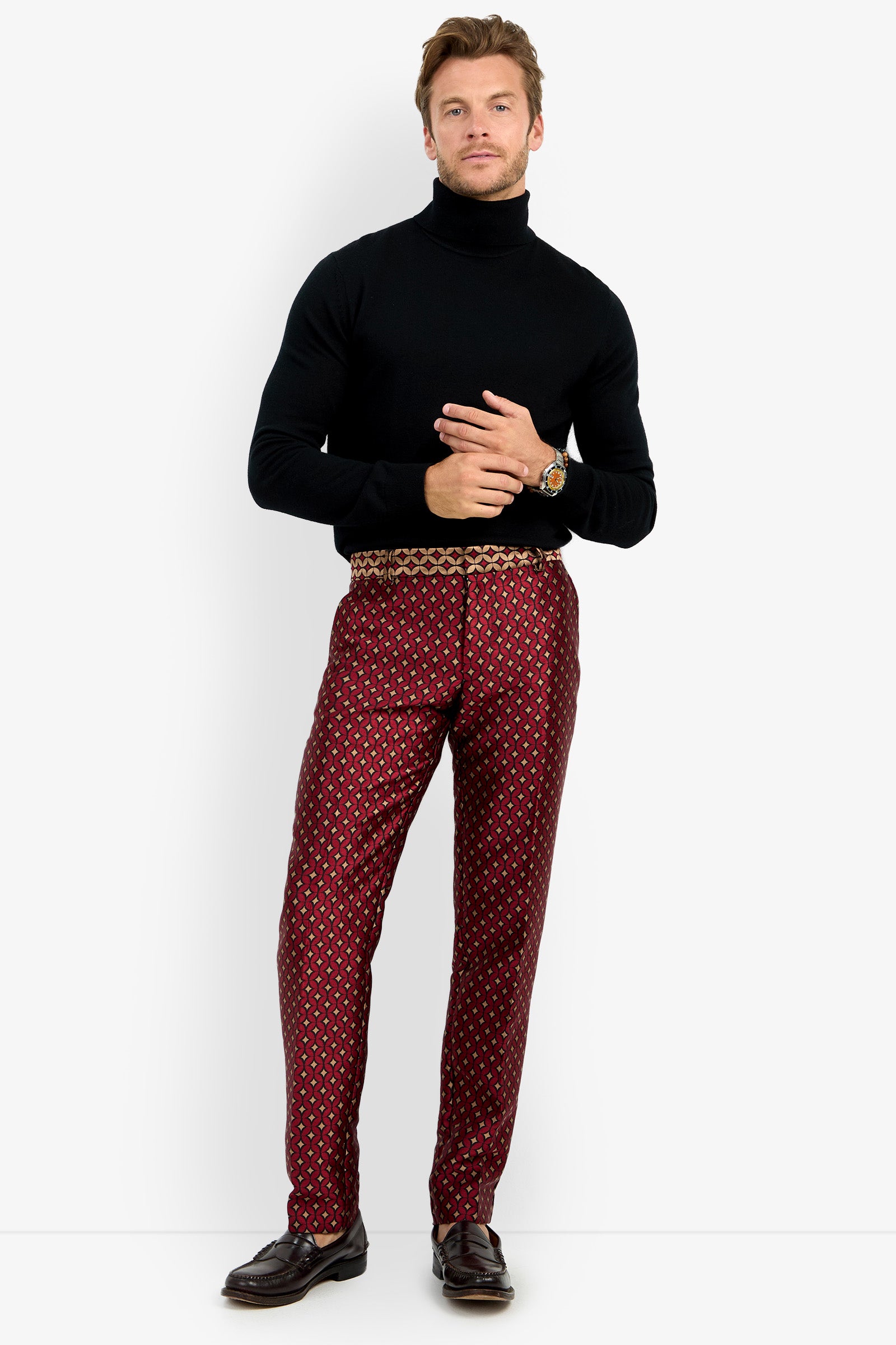 A man wearing a black turtleneck, CLYDE SLIM TROUSER patterned red pants, and black shoes stands against a white background, looking at the camera with a slight smile.