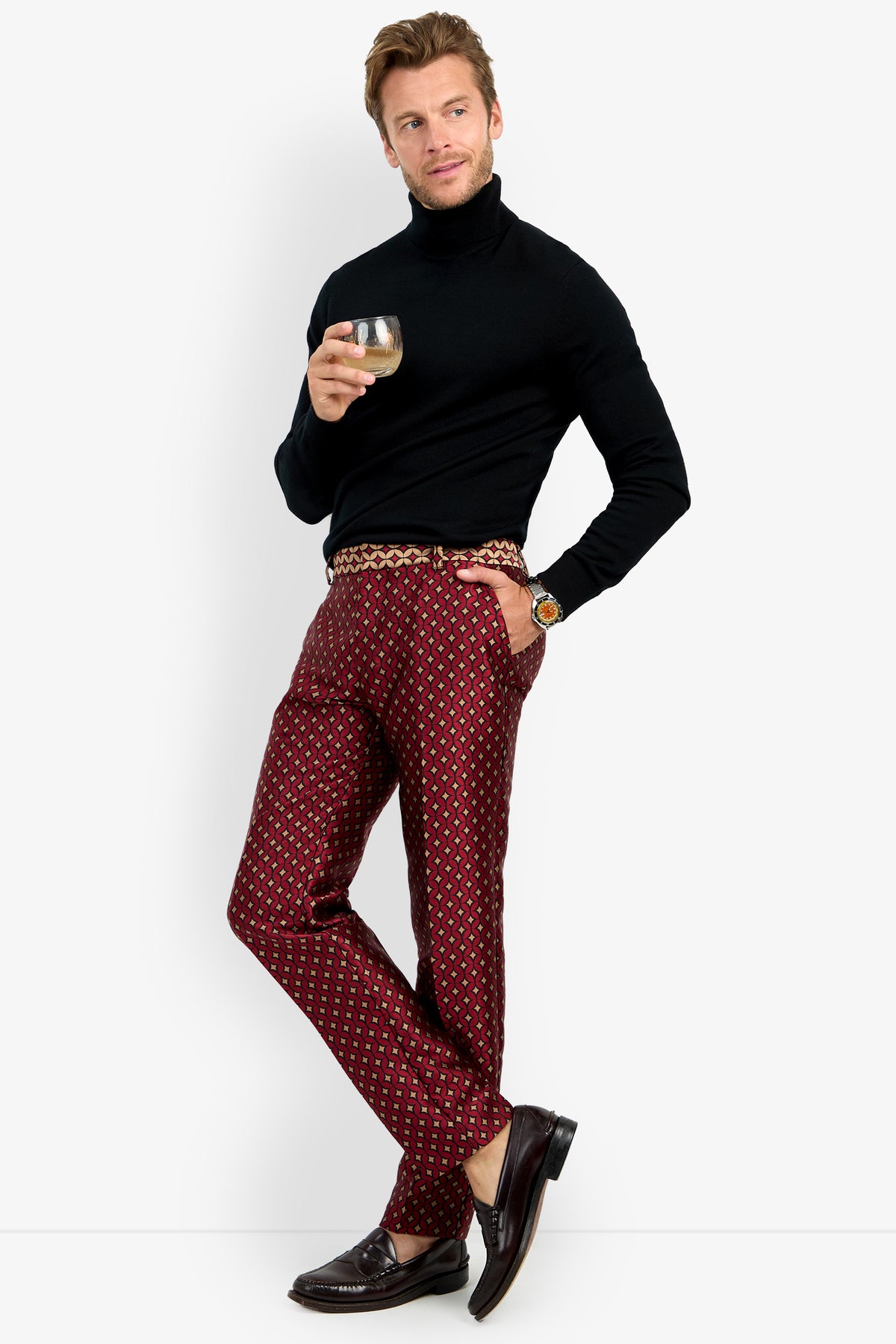 A man in a black turtleneck, CLYDE SLIM TROUSER in a red pattern, and brown loafers stands holding a drink. He has short light brown hair and wears a leopard print belt and watch, leaning casually against a white background.