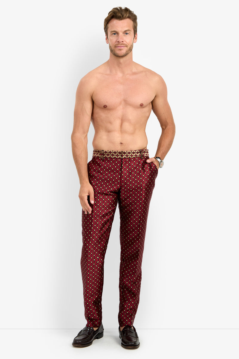 A shirtless man stands against a plain white background wearing the CLYDE SLIM TROUSER—red patterned pants—with his hands in his pockets, paired with black dress shoes and a watch.