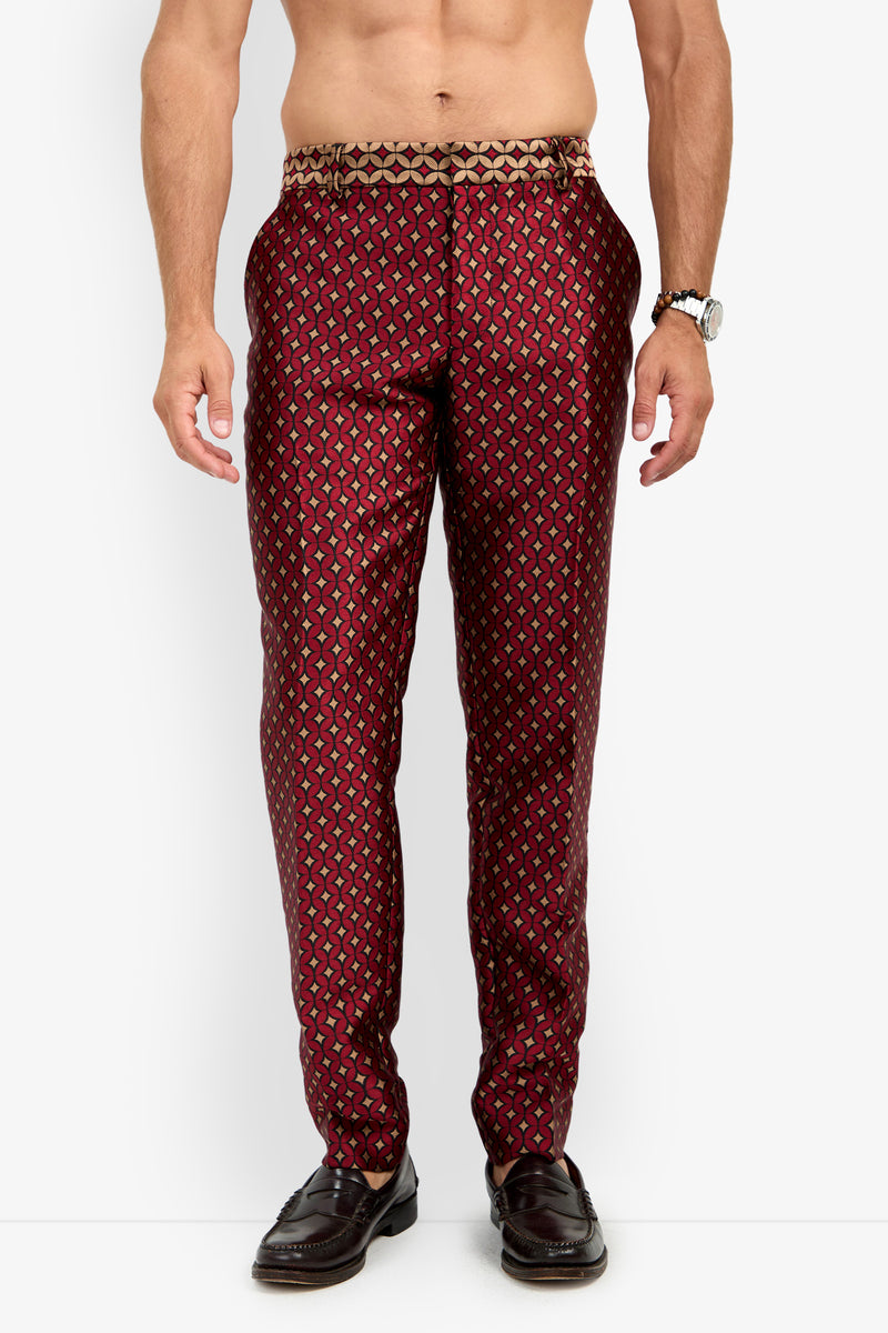 A shirtless man stands against a white background, wearing the CLYDE SLIM TROUSER in a red and gold pattern paired with brown loafers. His arms rest by his sides, and he has a watch on his left wrist.