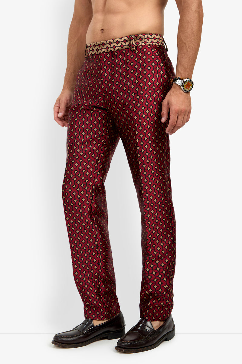 A man models the CLYDE SLIM TROUSER, featuring a maroon patterned design and a gold drawstring. He pairs the trousers with dark loafers and a wristwatch, standing bare-chested against a plain white background.