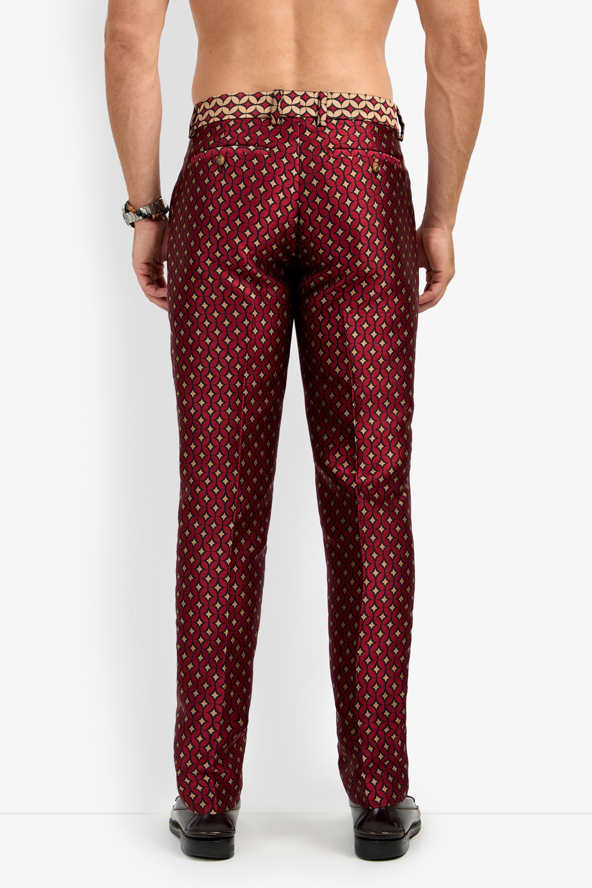 A man stands facing away, showing the back view while wearing the CLYDE SLIM TROUSER—shiny, patterned red and gold dress pants. He is shirtless and styled with black shoes, a beaded bracelet, and a ring on his right hand.