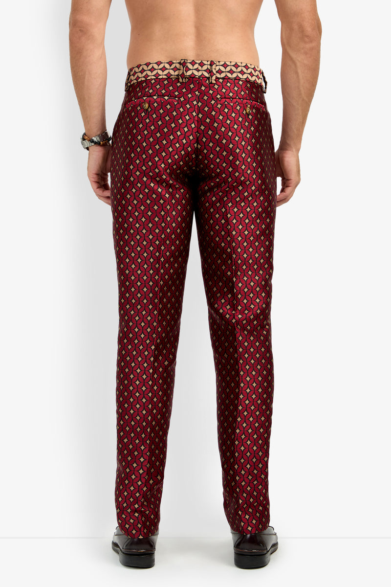A man stands facing away, showing the back view while wearing the CLYDE SLIM TROUSER—shiny, patterned red and gold dress pants. He is shirtless and styled with black shoes, a beaded bracelet, and a ring on his right hand.