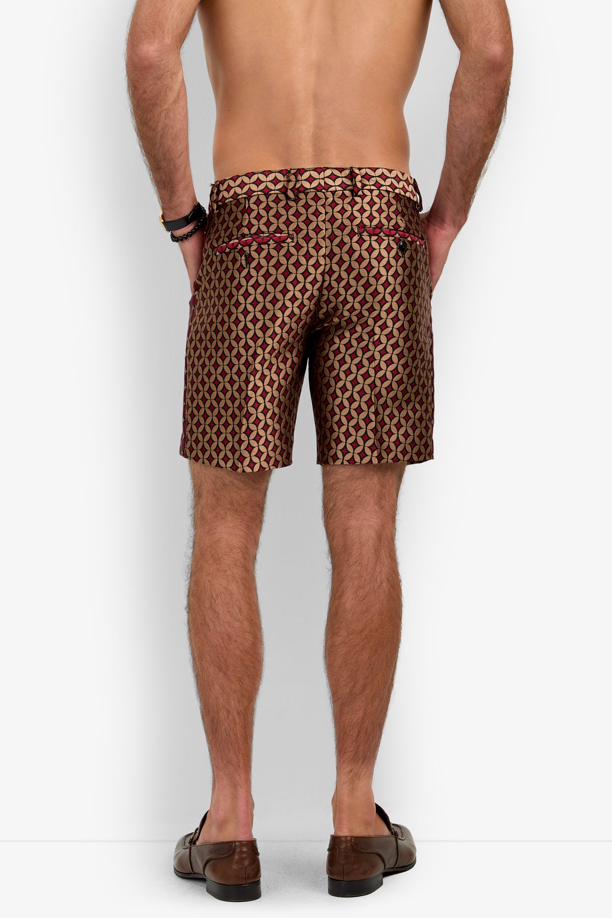 A man stands facing away, shirtless, wearing the Cataline Short—patterned in maroon, gold, and black—paired with brown loafers and black bracelets against a plain white background.