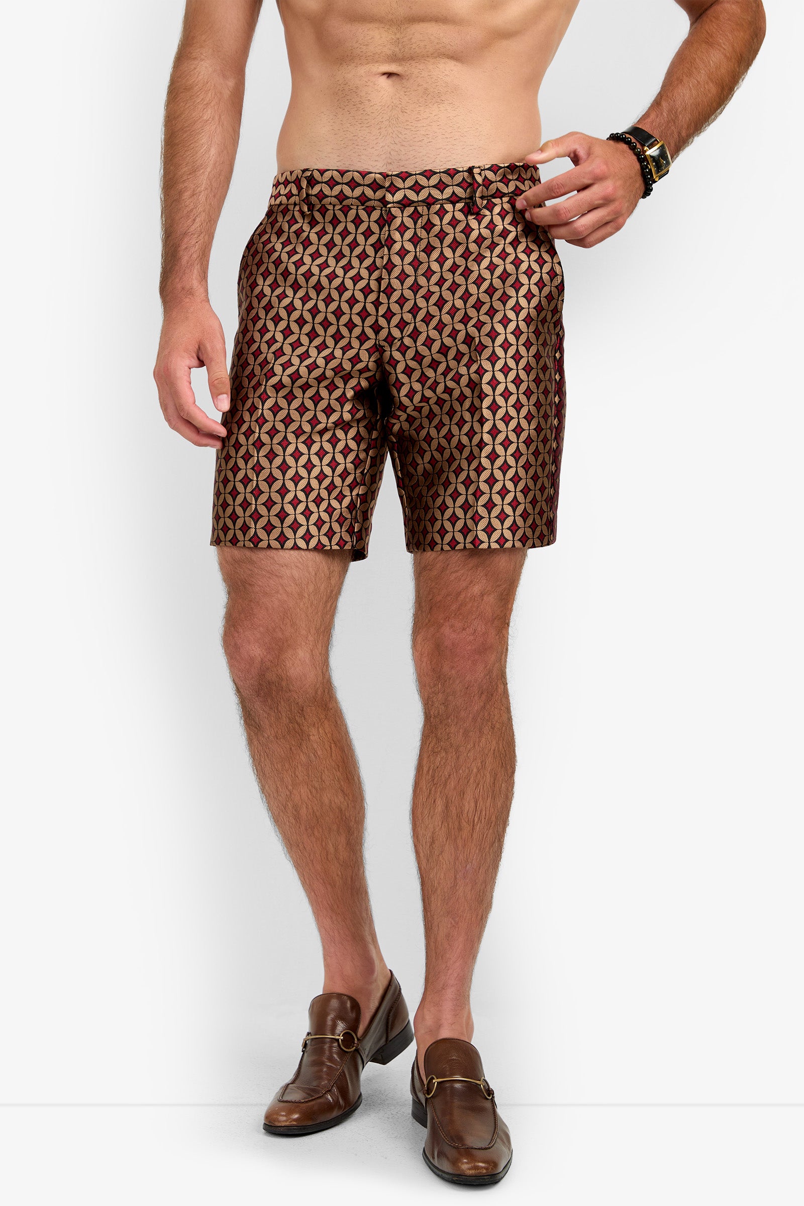 A shirtless man stands against a white background wearing the Cataline Short—patterned brown shorts paired with brown loafers, his hand in one pocket and the other by his side.