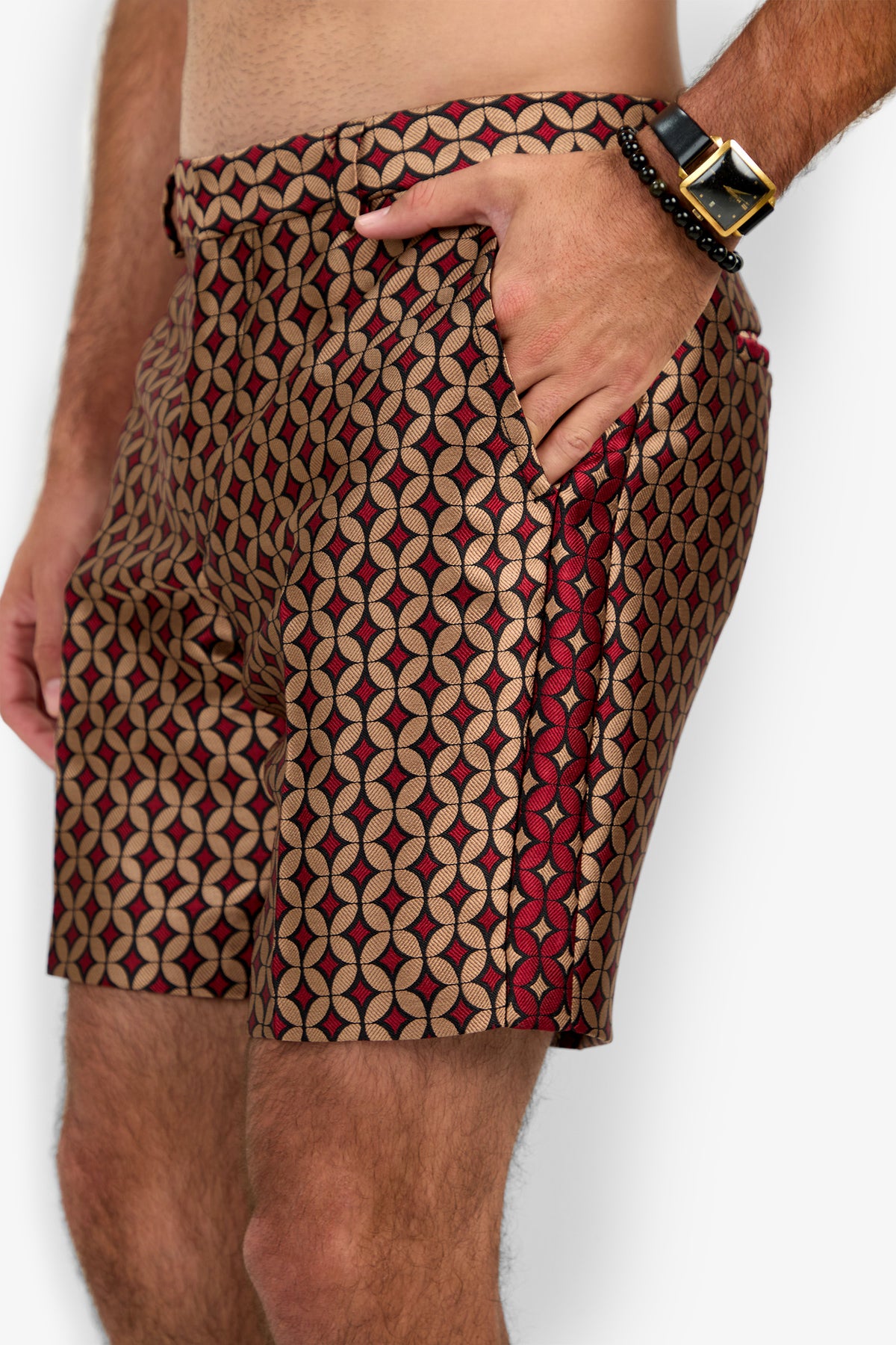 A person wears the Cataline Short, featuring red, beige, and black geometric patterns, and stands with one hand in their pocket, displaying a watch and beaded bracelet on their wrist.