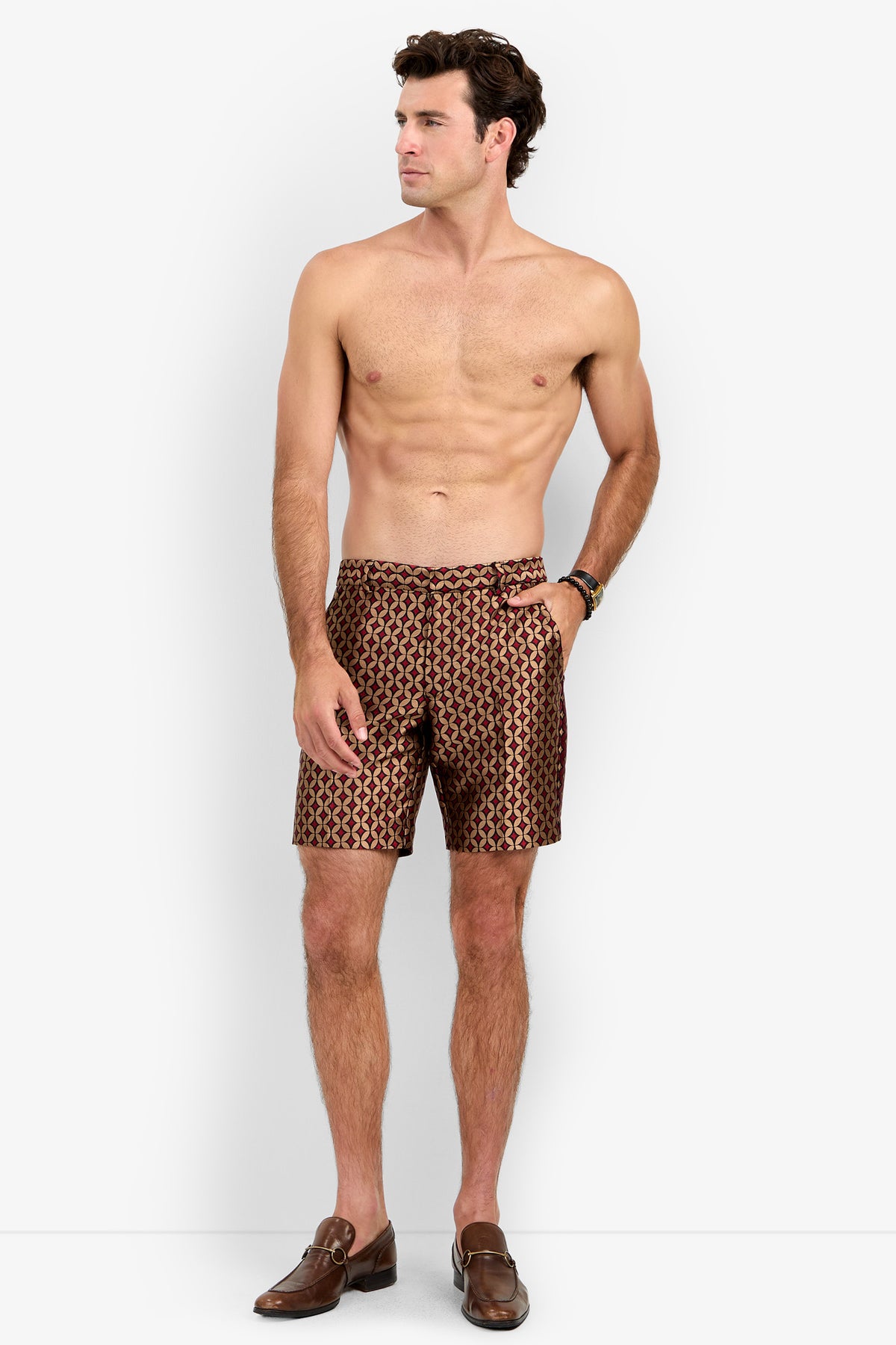 A shirtless man stands facing left, wearing the patterned brown Cataline Short and brown loafers, with one hand in his pocket against a plain white background.