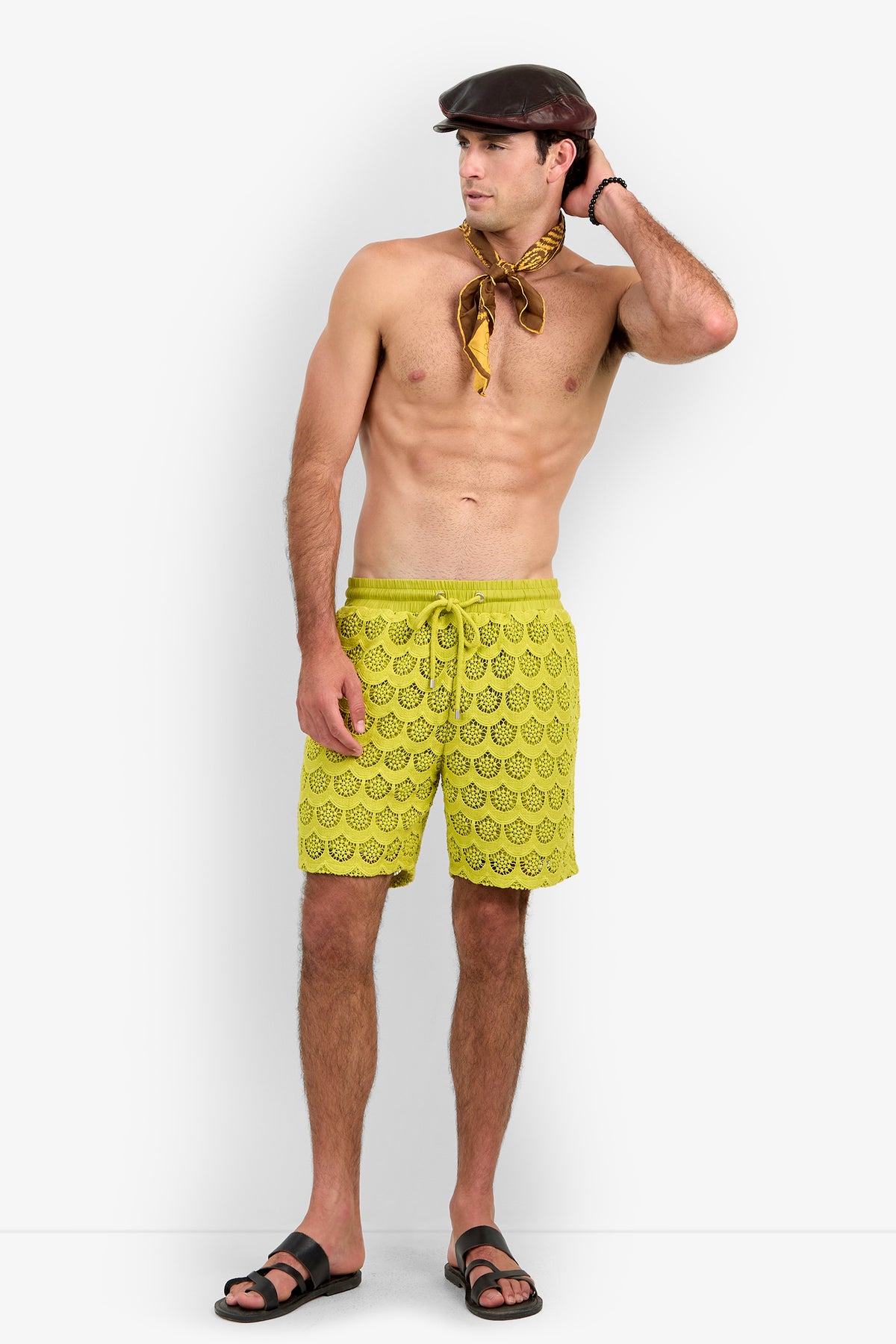 A shirtless man wearing the Shoreline Short in yellow, paired with black sandals, a brown flat cap, and a yellow scarf around his neck, stands with one hand on his head looking to the side against a plain white background.