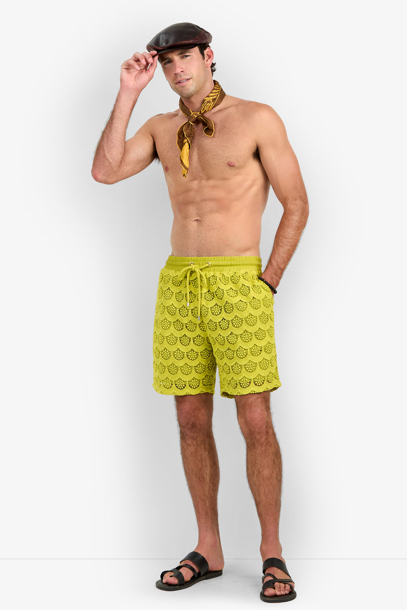 A man wearing a brown cap, yellow scarf, Shoreline Short swim trunks with a yellow pattern, and black sandals stands against a white background with one hand touching his cap and the other in his pocket.