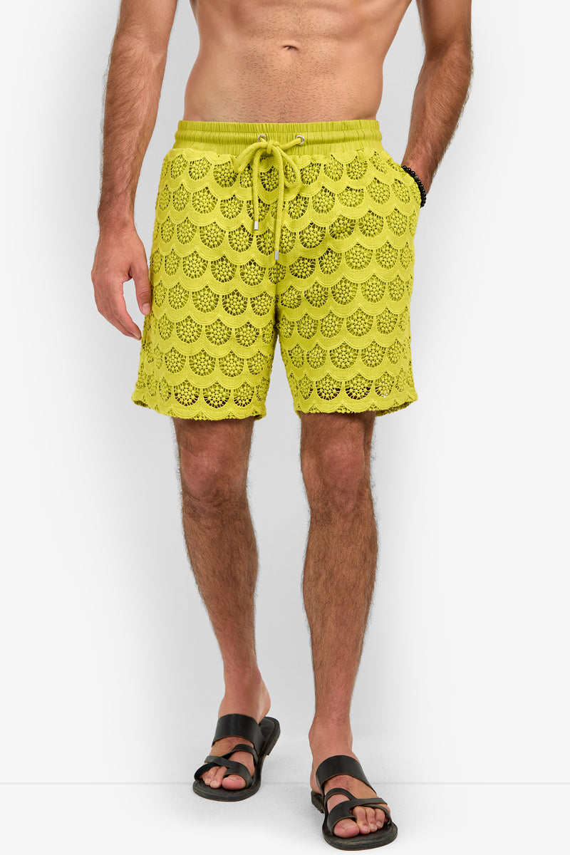 A person wearing the Shoreline Short in yellow-green, featuring scalloped crochet patterns and a drawstring waist, paired with black sandals and standing against a white background.
