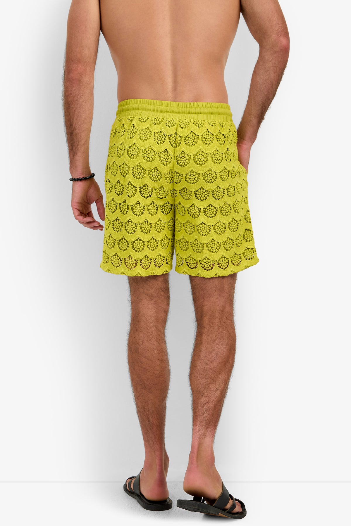 A man stands facing away, wearing the Shoreline Short in yellow crochet, paired with black sandals and a black wristband against a plain white background.