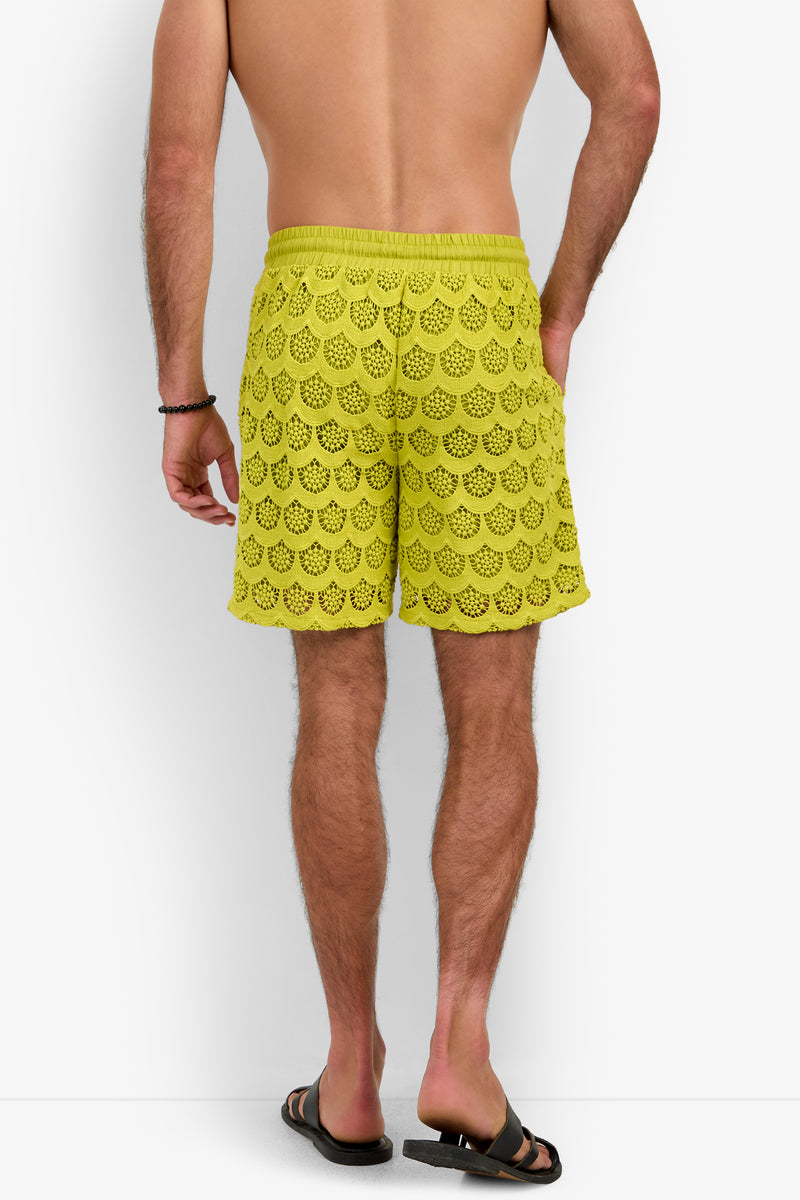 A man stands facing away, wearing the Shoreline Short in yellow crochet, paired with black sandals and a black wristband against a plain white background.