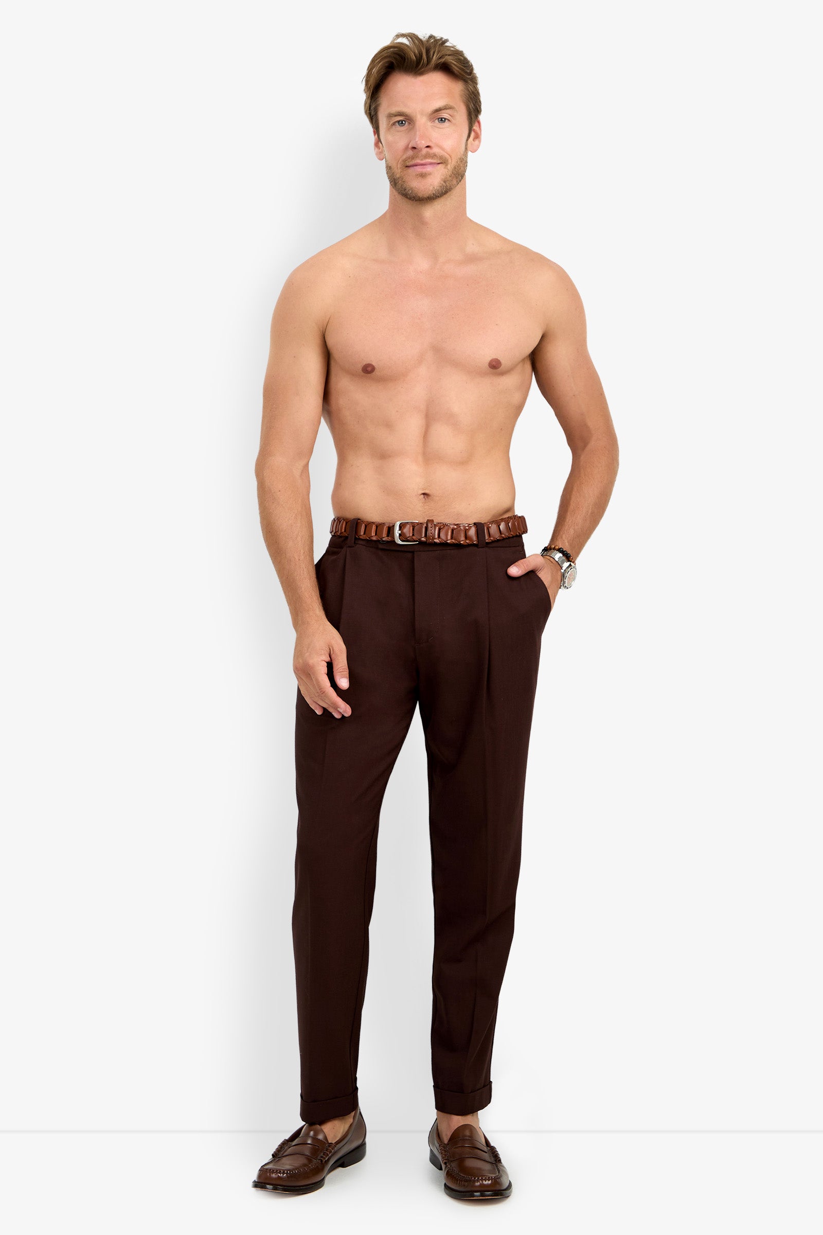 A shirtless man stands and smiles against a plain white background, wearing the BEALE PLEATED TROUSER paired with a brown belt and brown loafers.