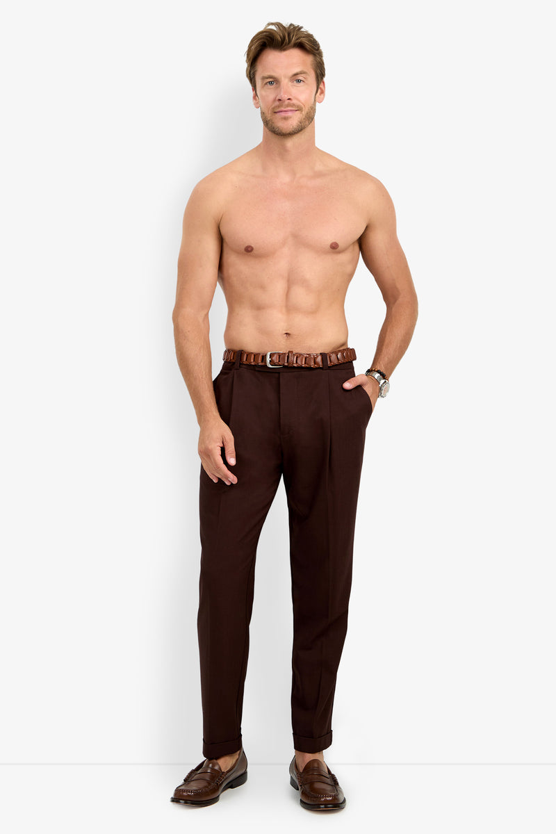 A shirtless man stands and smiles against a plain white background, wearing the BEALE PLEATED TROUSER paired with a brown belt and brown loafers.