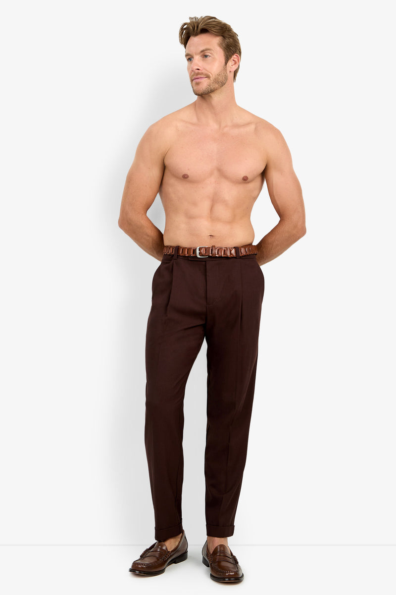 A shirtless man with short hair wears the BEALE PLEATED TROUSER in dark brown, paired with a matching belt and brown loafers, standing against a plain white background with his hands in his pockets.