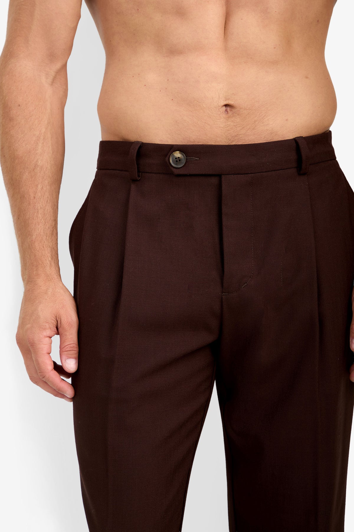 A shirtless person wearing the BEALE PLEATED TROUSER—high-waisted dark brown dress pants with a button and belt loops—stands against a plain background.