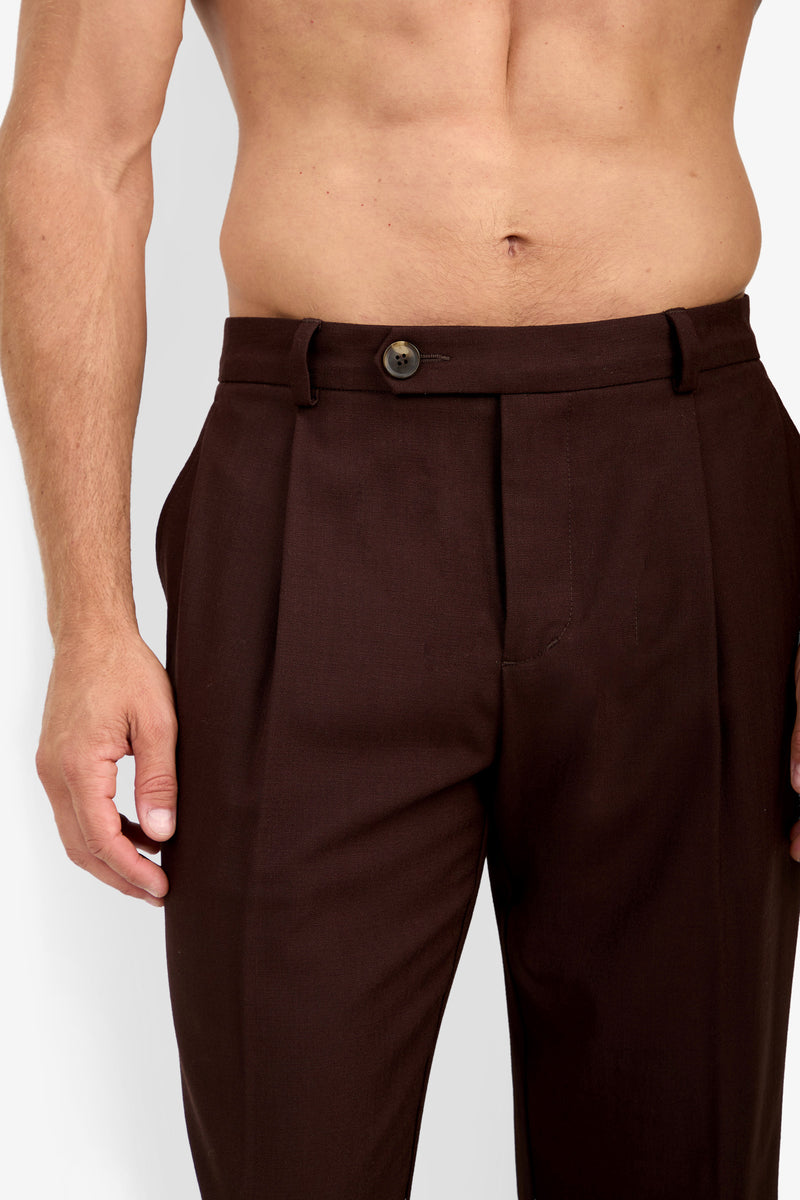 A shirtless person wearing the BEALE PLEATED TROUSER—high-waisted dark brown dress pants with a button and belt loops—stands against a plain background.