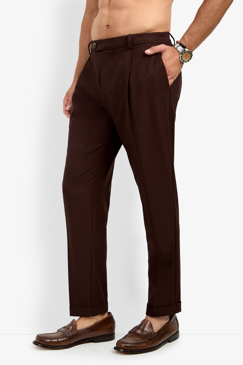 A man wearing the BEALE PLEATED TROUSER in dark brown with brown loafers, showing only his torso and legs. He has a watch on his left wrist and his hands are in his pockets.