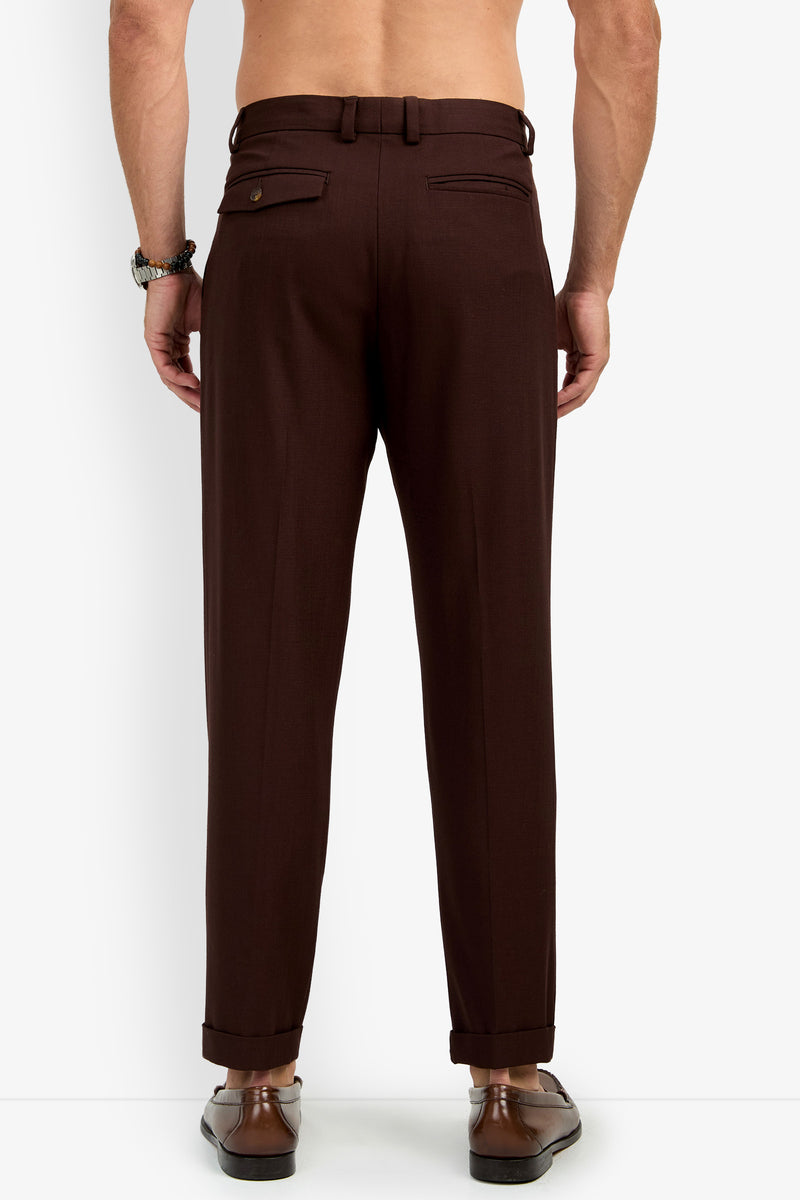 A shirtless man, seen from the back, wears the BEALE PLEATED TROUSER in brown with cuffs, paired with brown dress shoes. He has a bracelet on his left wrist. The trousers feature two back pockets with buttons.