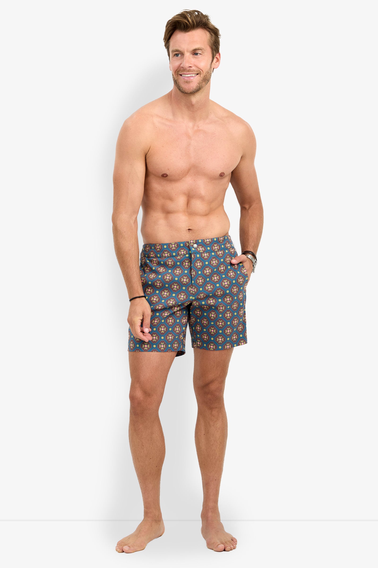 A man with short hair stands barefoot and smiling, shirtless, wearing the HONOLULU BOARDSHORT featuring a blue pattern with red and yellow accents, against a plain white background.