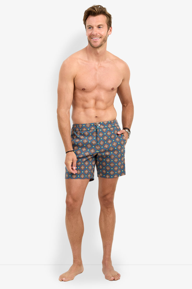 A man with short hair stands barefoot and smiling, shirtless, wearing the HONOLULU BOARDSHORT featuring a blue pattern with red and yellow accents, against a plain white background.