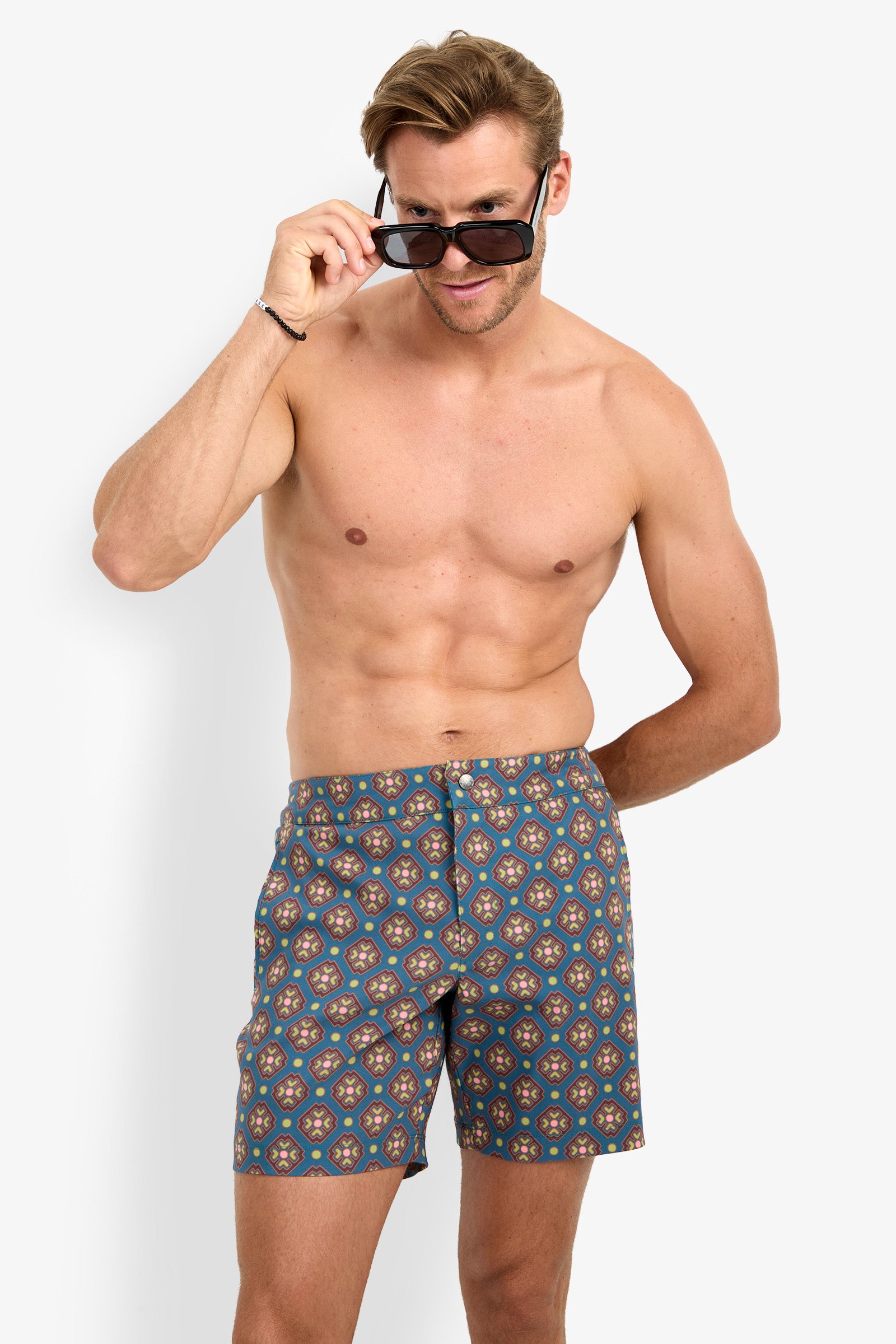 A shirtless man models the HONOLULU BOARDSHORT swim trunks, looking at the camera with a smile as he lowers his sunglasses and places one hand behind his back, set against a plain white background.