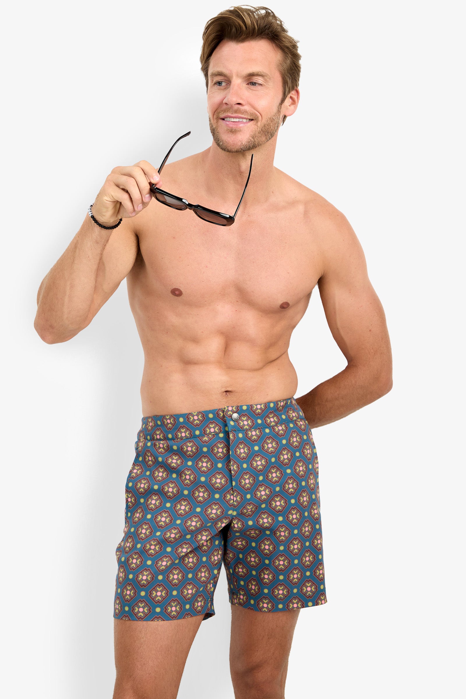 A shirtless man with light brown hair smiles while holding sunglasses, wearing the HONOLULU BOARDSHORT featuring a geometric blue pattern, standing against a plain white background.