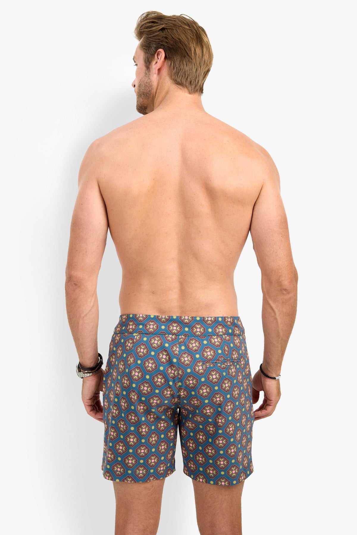 A shirtless man with light brown hair stands facing away against a plain white background, wearing the HONOLULU BOARDSHORT featuring a blue pattern. His arms are relaxed at his sides.