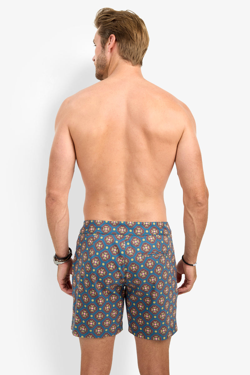 A shirtless man with light brown hair stands facing away against a plain white background, wearing the HONOLULU BOARDSHORT featuring a blue pattern. His arms are relaxed at his sides.