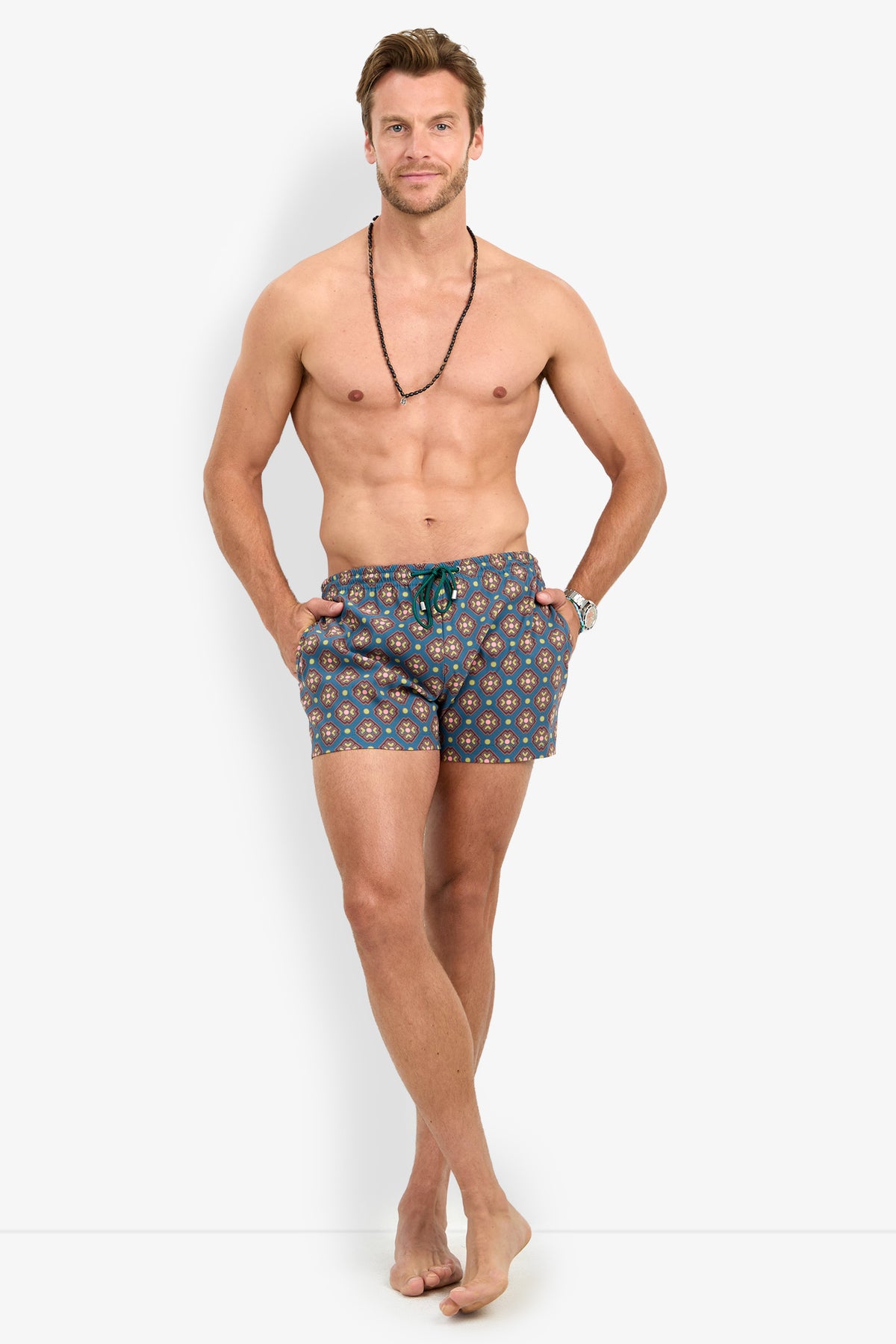 A shirtless man smiles at the camera against a plain white background, barefoot with his hands in his pockets, wearing the OCEANSIDE SWIM TRUNK and a necklace.