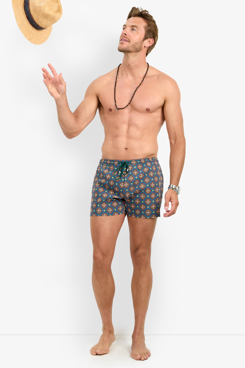 A shirtless man models the OCEANSIDE SWIM TRUNK and a beaded necklace, tossing a straw hat in the air. He stands barefoot against a plain white background, looking up at the hat with a relaxed expression.