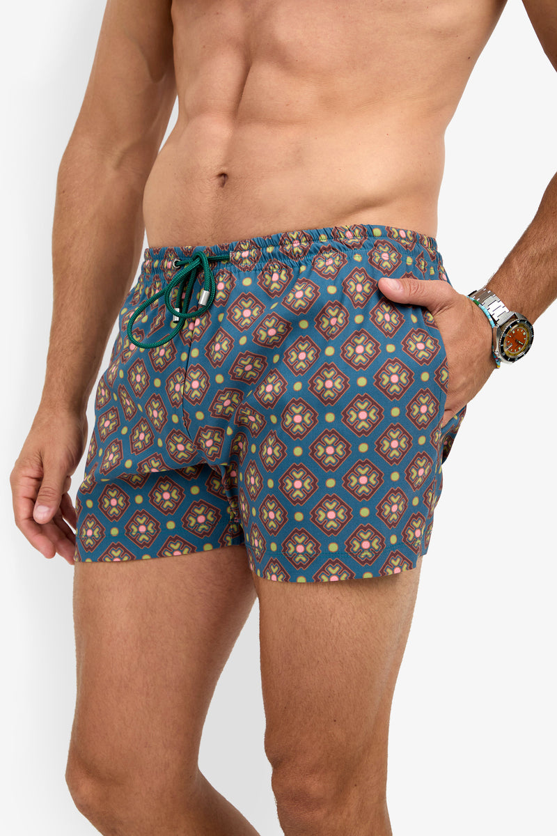 A man wearing the OCEANSIDE SWIM TRUNK, featuring a blue and multicolored geometric design, stands with one hand in his pocket and a watch on his wrist.