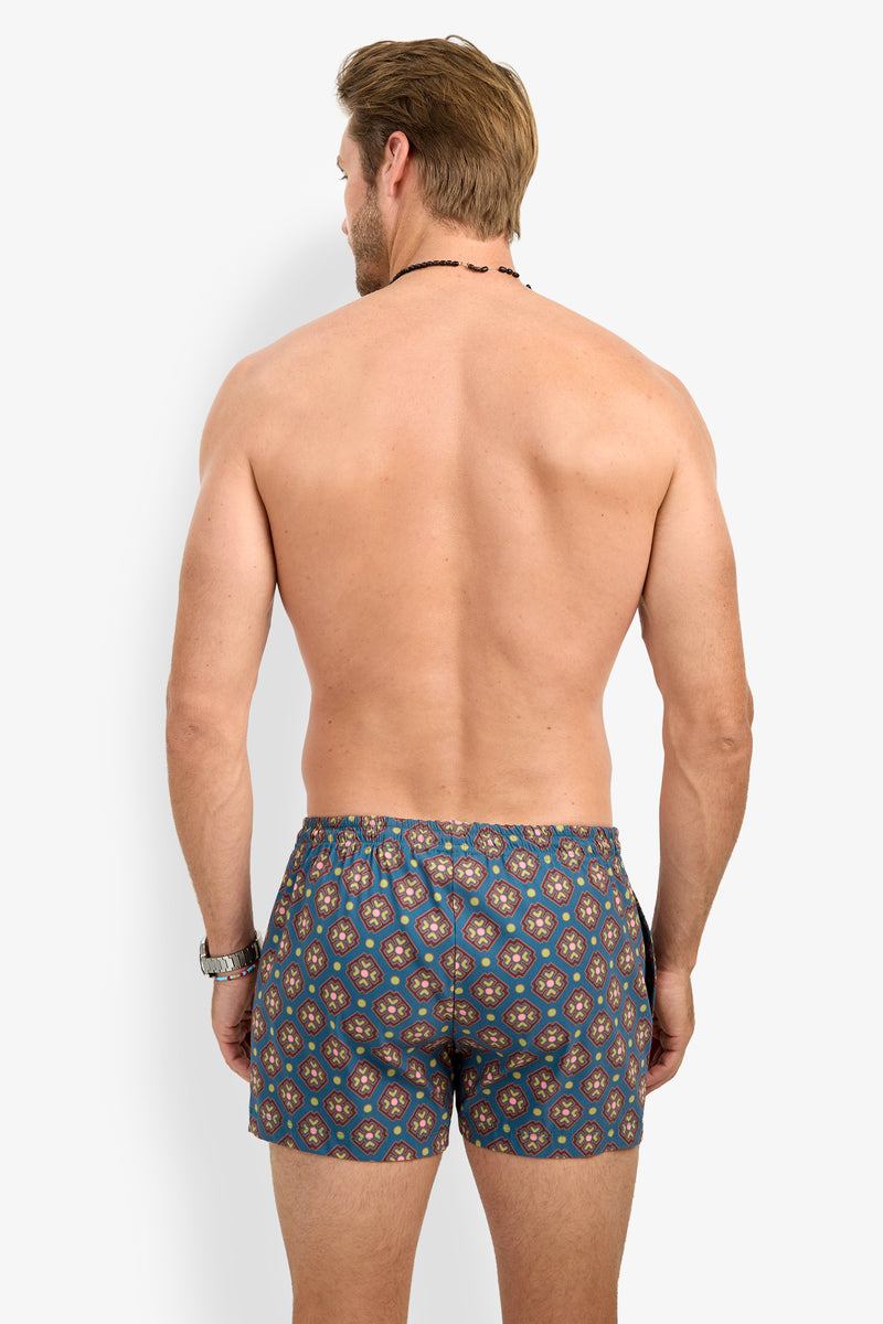 A shirtless man with light brown hair stands facing away, wearing the OCEANSIDE SWIM TRUNK—patterned blue shorts—along with a black beaded necklace. A watch is on his left wrist, and his arms rest relaxed by his sides.