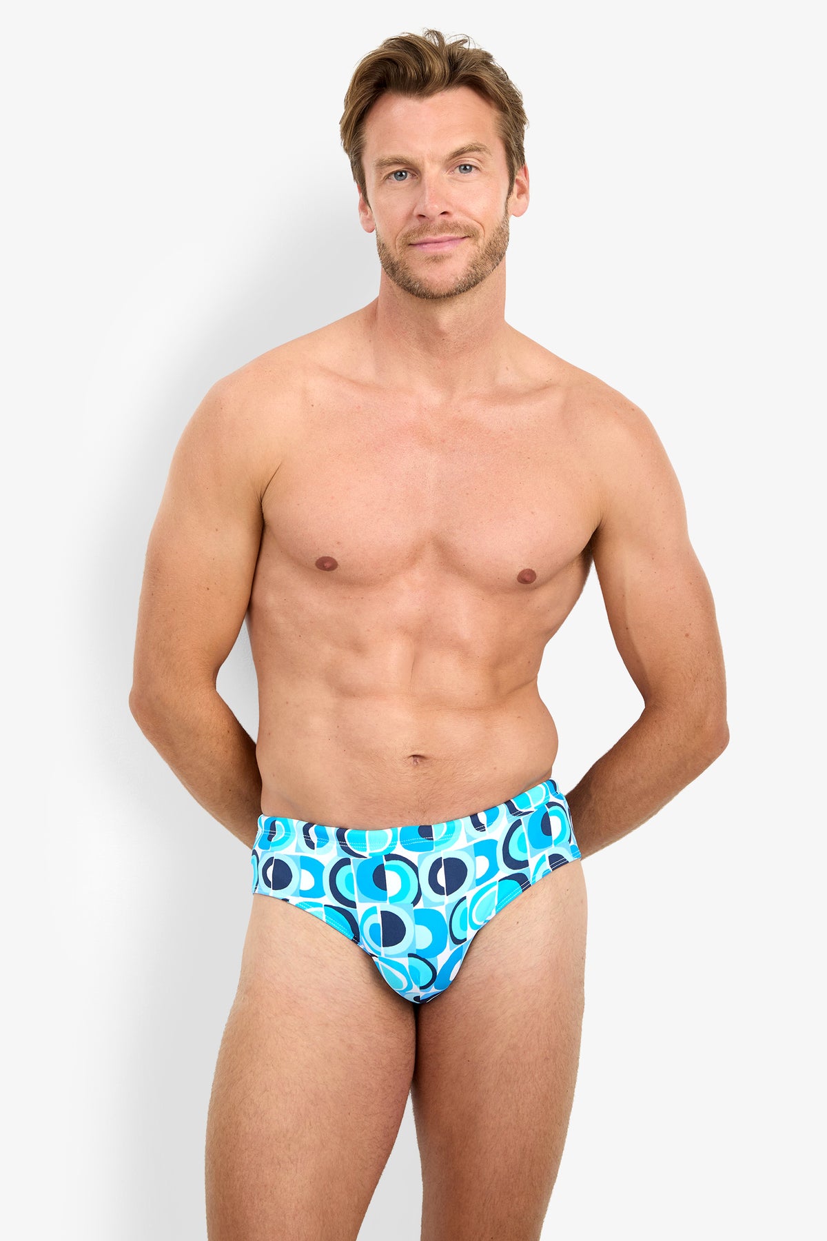 A fit, smiling man stands against a white background, wearing the BONDI SWIM BRIEF, featuring a blue pattern with white, dark blue, and teal geometric designs.