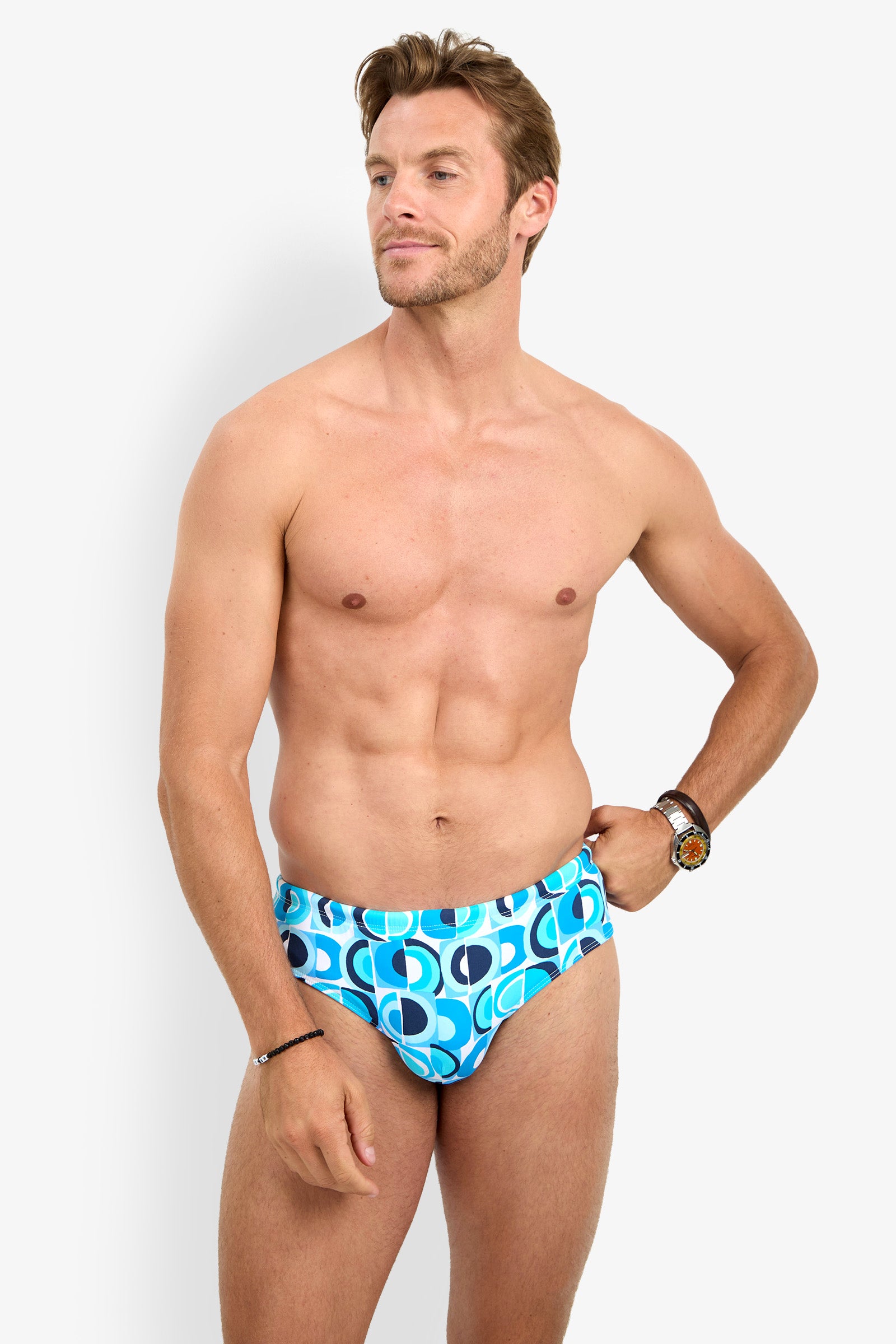 A man stands against a white background wearing the BONDI SWIM BRIEF, a watch, and a bracelet. He is looking to the side with one hand resting on his hip.